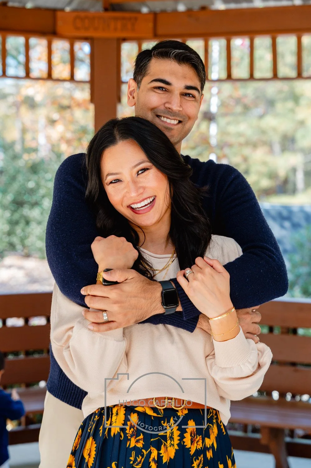 A smiling couple hugging outdoors in a park pavilion with autumn trees in the background.