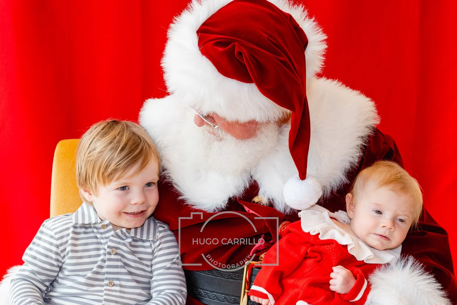 Santa Claus with white beard in red and white outfit sitting with two young children, one smiling and one looking thoughtful, in front of a red backdrop.