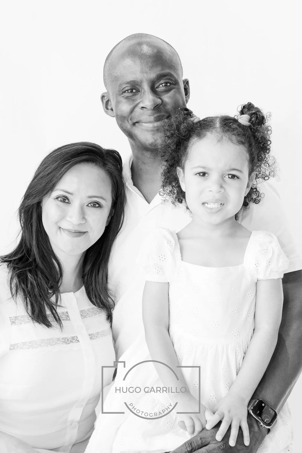 A portrait of a diverse family of three, smiling and posing together against a plain white background. The woman has long dark hair, the man is bald with a broad smile, and the young girl has curly hair styled in pigtails and is making a slightly con