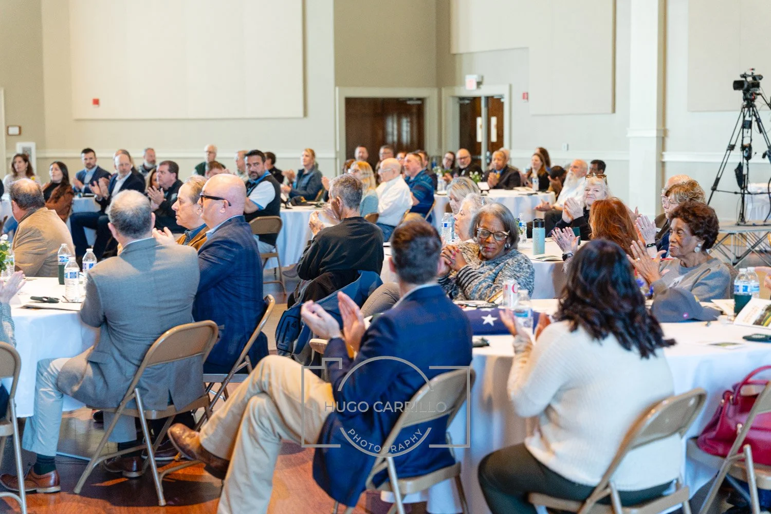 A diverse group of people seated at round tables in a conference room, listening and applauding during a formal event or presentation.