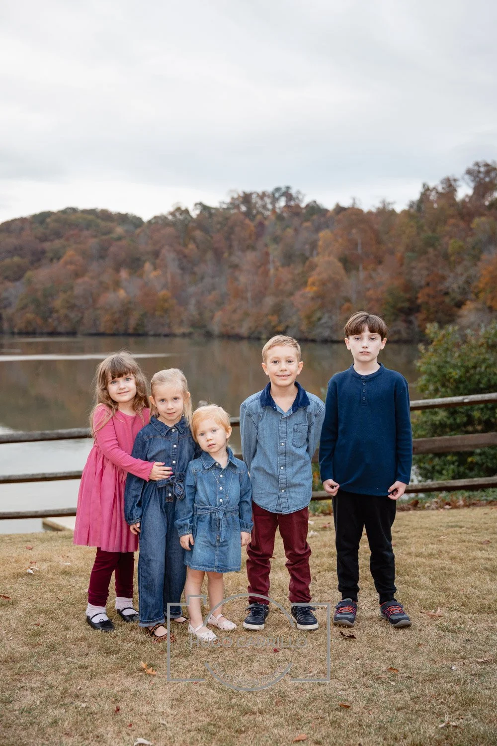 Group of five children standing outdoors near a lake with autumn trees in the background.
