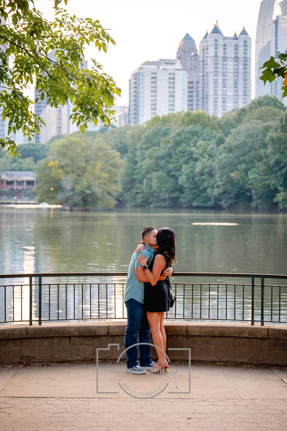 A couple kissing on a railing in a park by a lake, with trees and city high-rise buildings in the background.
