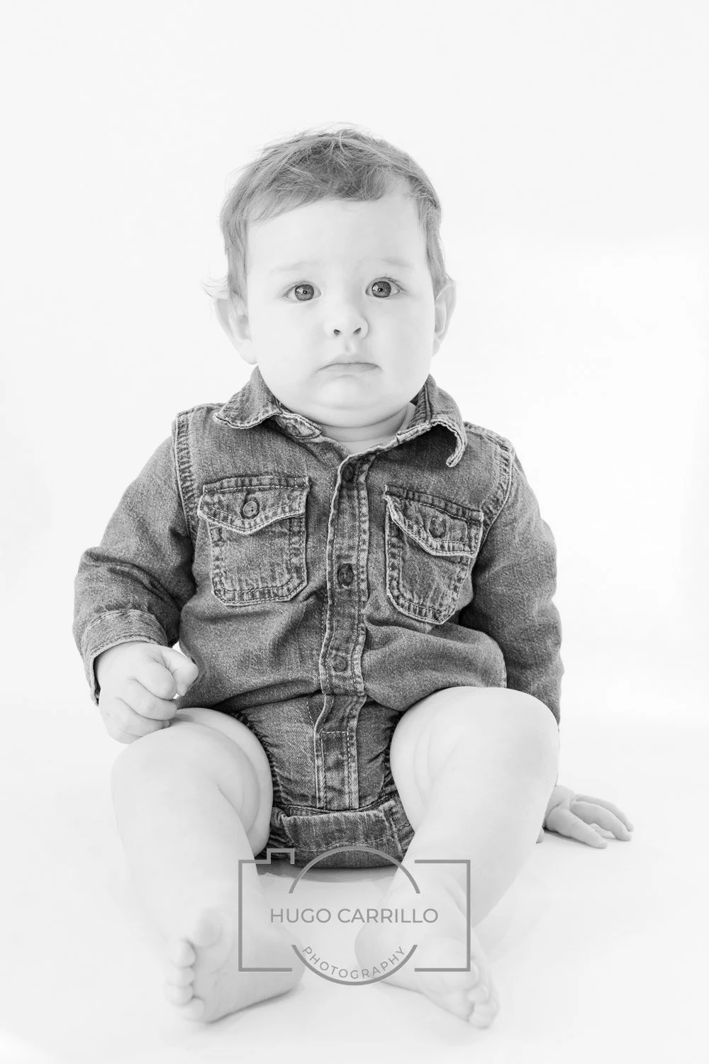 Black and white photo of a young child sitting on the floor against a white background, wearing a denim jacket, with a serious expression.