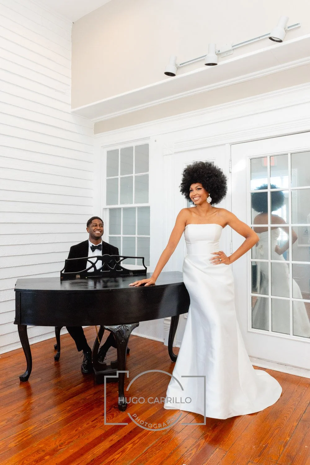 Woman in a white strapless gown standing next to a black grand piano with a man in a tuxedo sitting at the piano. The setting is a white room with large glass doors and hardwood floors.