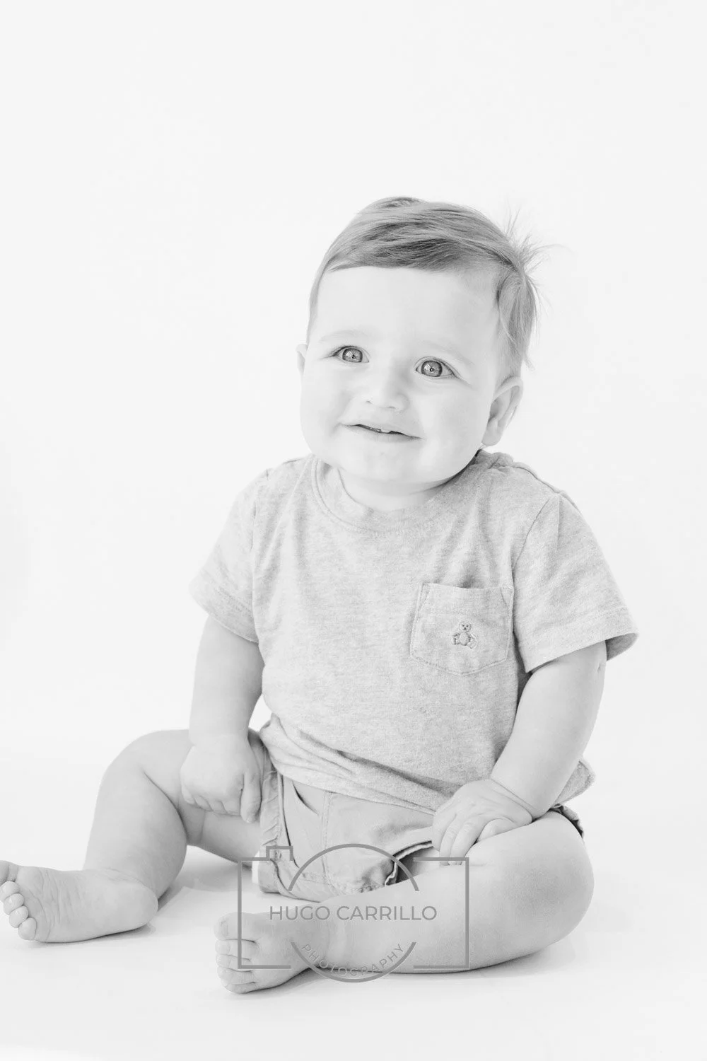 A young toddler with light hair and light eyes, sitting on the floor against a plain white background, smiling and looking slightly to the right, wearing a short-sleeved gray T-shirt and shorts. The photo has a watermark from Hugo Carrillo Photograph
