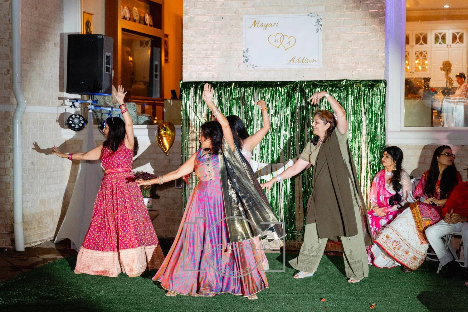 Group of women dancing at a celebration event, dressed in colorful traditional Indian attire, with a decorated backdrop and seated guests observing.