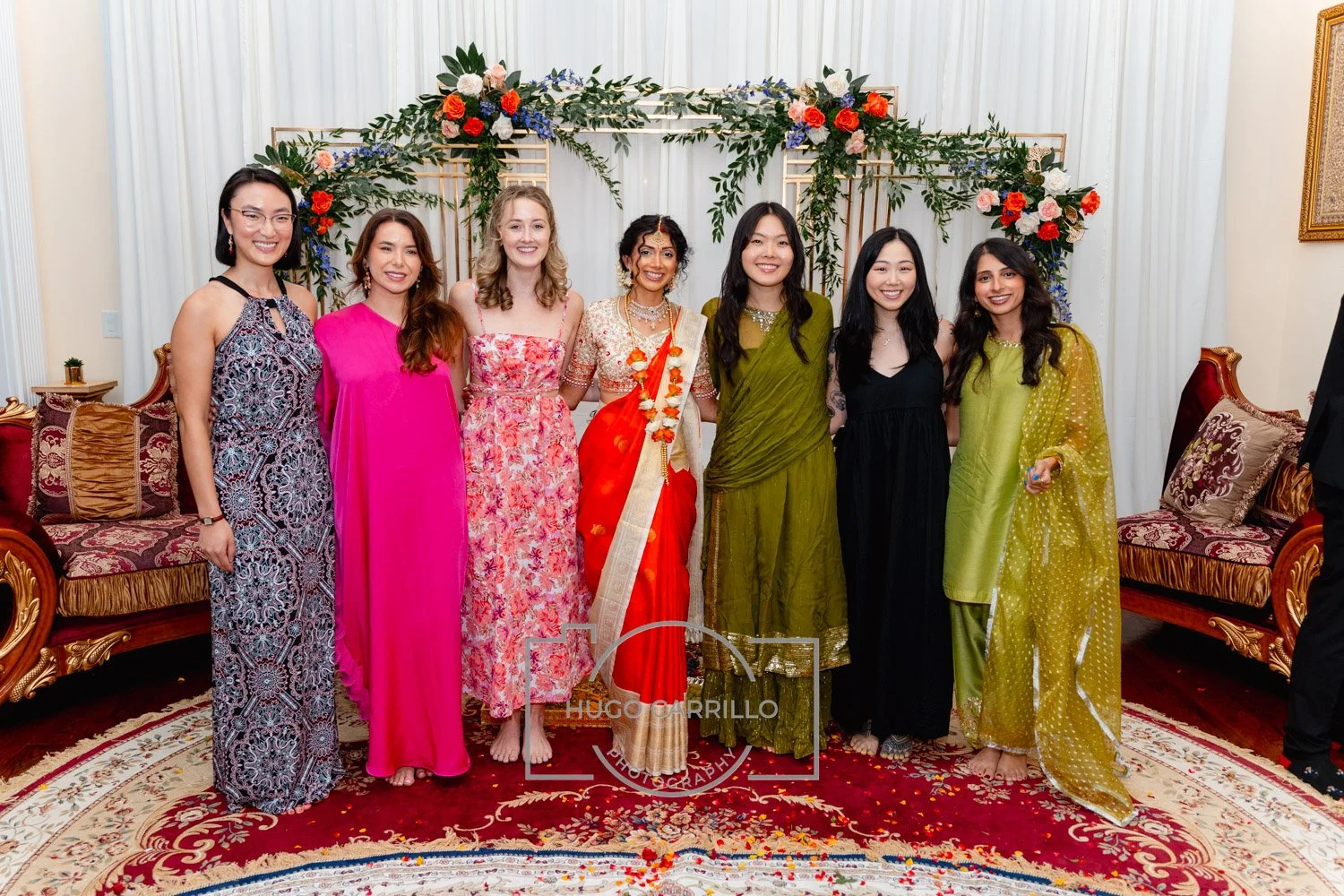 Group of eight women in colorful traditional and formal dresses standing in a decorated room with floral arrangements on the background.
