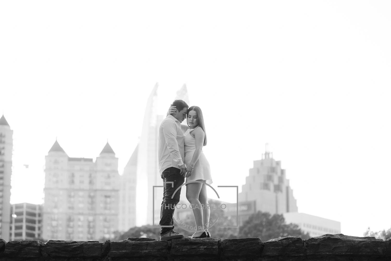 A black-and-white photo of a young man and woman standing close together on a stone surface, with a city skyline featuring tall buildings and a unique.Ferris wheel in the background.