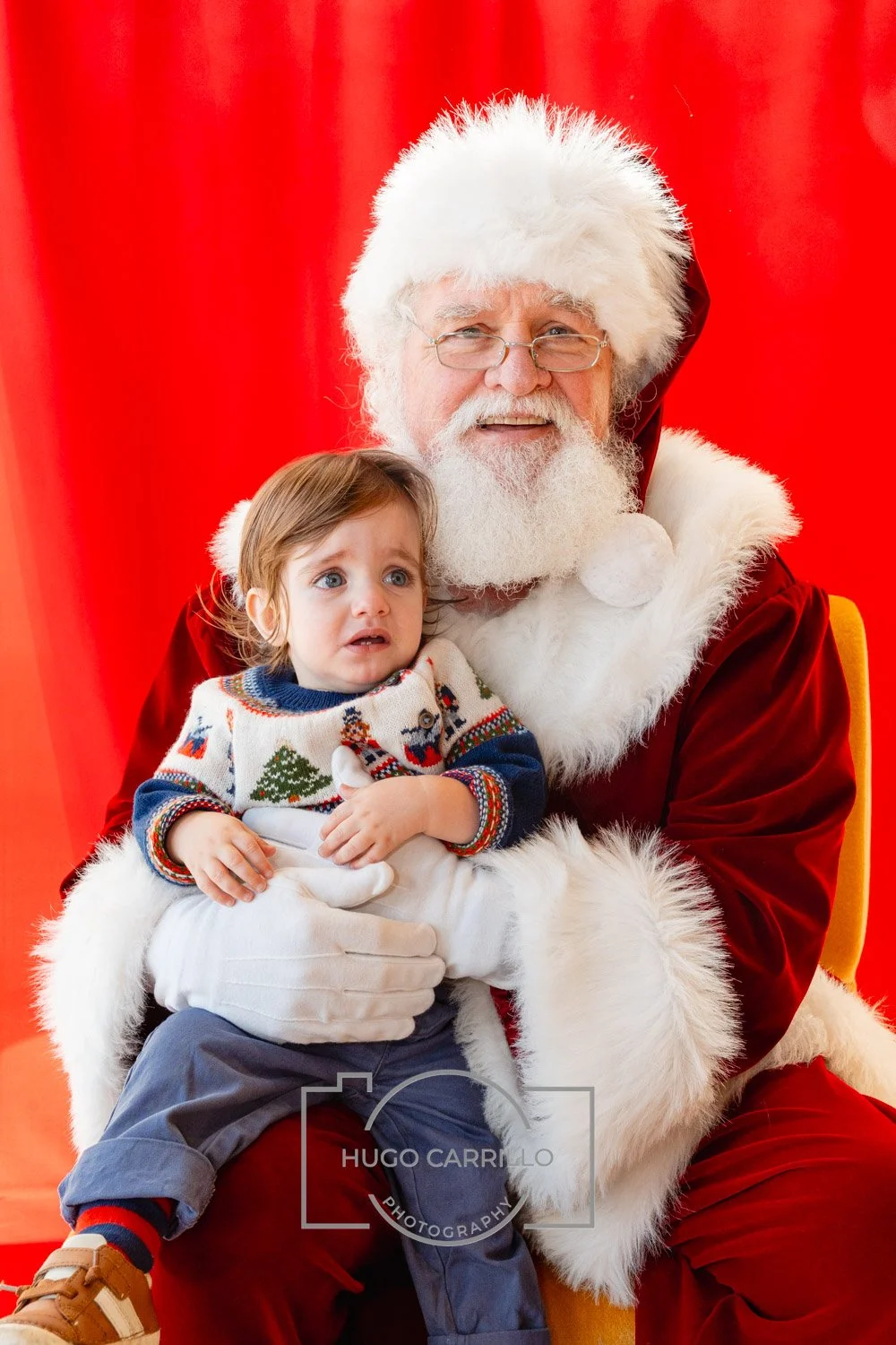 A young boy with a distressed expression sitting on Santa Claus's lap, with a red background.