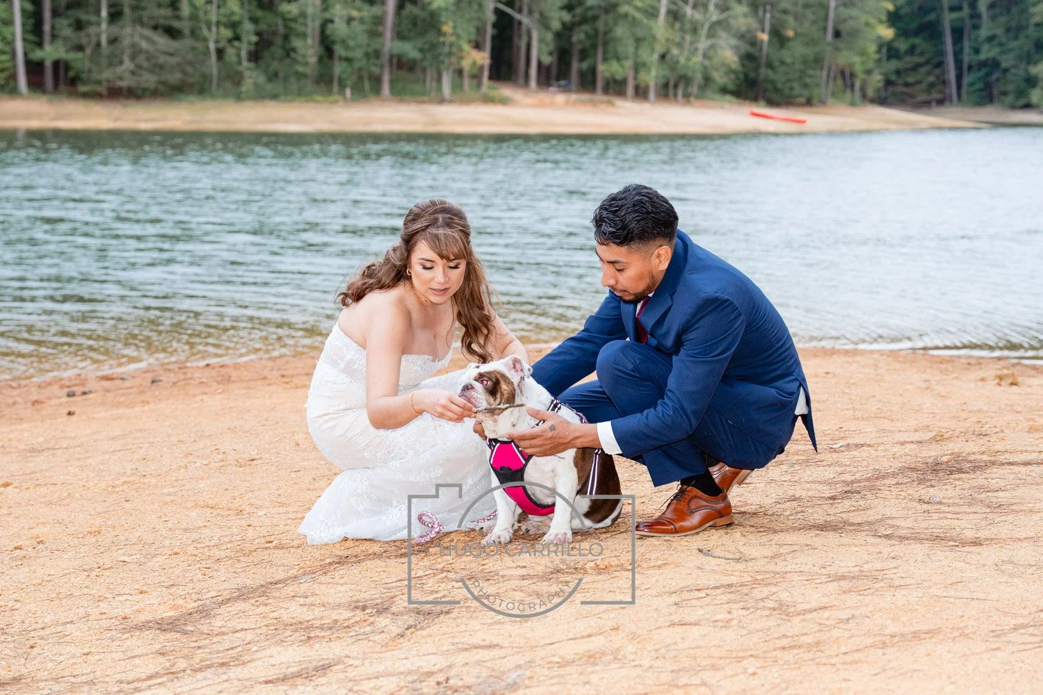 A couple dressed in wedding attire kneeling on a sandy beach by a lake with an English bulldog. The woman is wearing a white strapless wedding gown and has long brown hair. The man is in a blue suit and brown shoes. The woman is holding the dog's fac