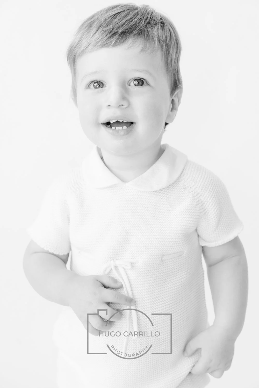 Black and white portrait of a young boy with short hair, smiling with an open mouth, wearing a textured short-sleeve shirt, standing against a plain background.