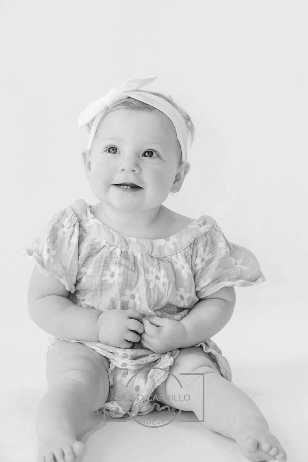 Black and white photo of a smiling baby girl with a bow headband, wearing a patterned dress, sitting on a white surface.