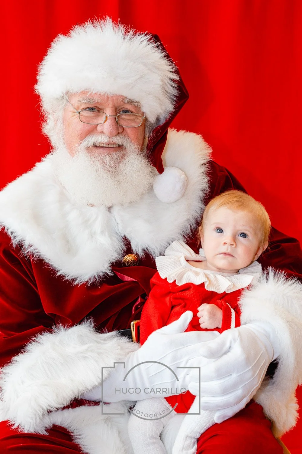A man dressed as Santa Claus holding a young girl dressed in a red Christmas outfit, both against a red background.