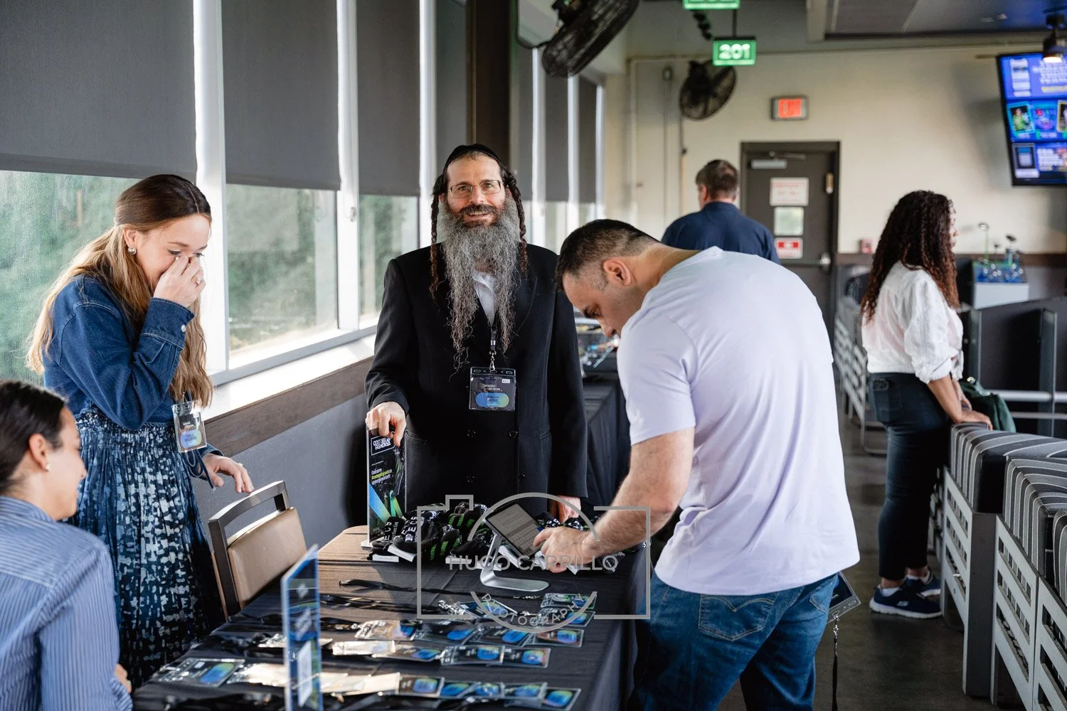 People browsing tech gadgets and accessories on a table at an indoor event, with a man with a long beard and a suit standing behind the table, smiling.