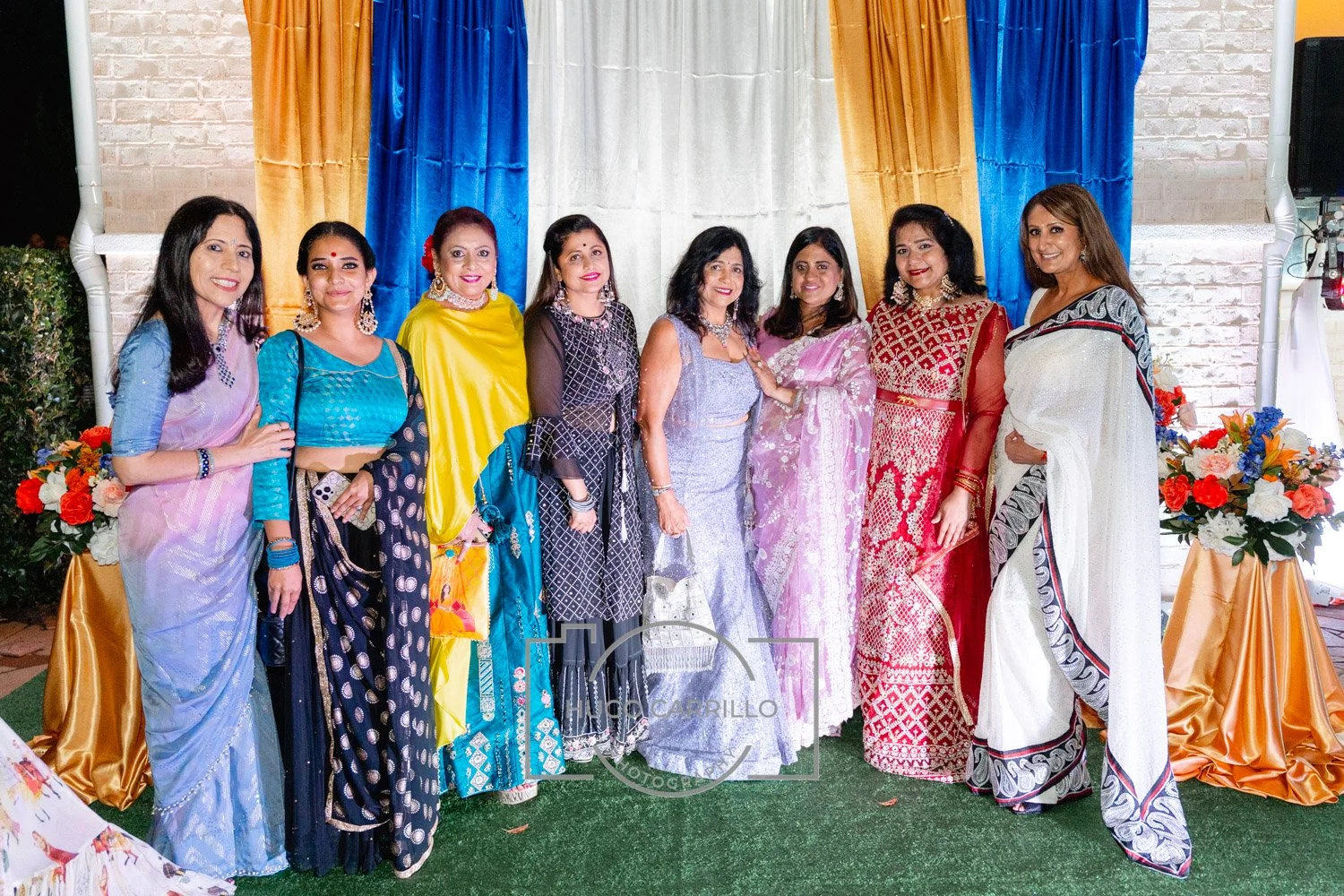 A group of ten women dressed in colorful sarees and traditional Indian attire standing in front of a decorative backdrop with blue, white, and yellow curtains, and floral arrangements.