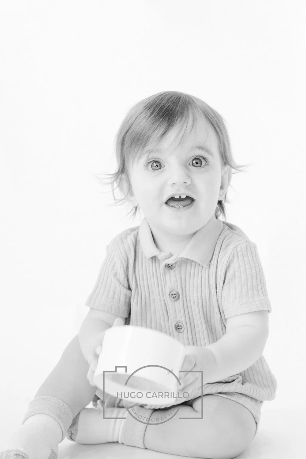 Black and white photo of a young child with wide eyes and an open mouth, sitting cross-legged and holding a mug, wearing a short-sleeved, button-up shirt.