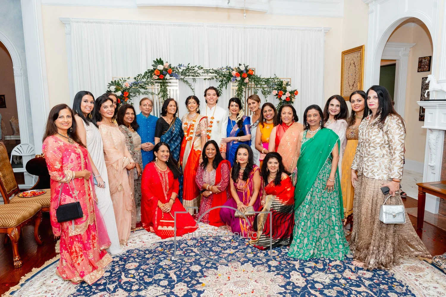 Group of women and men dressed in colorful traditional Indian attire, posing together at a celebration or wedding, standing on a decorative rug in front of a floral backdrop.