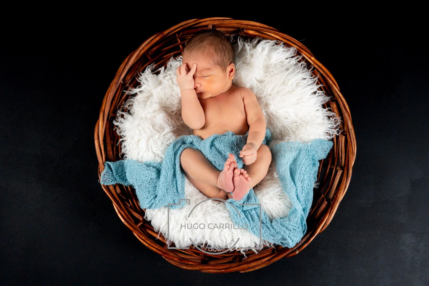 Sleepy newborn baby lying in a round wicker basket on a fluffy white blanket with a blue knitted cloth, black background.