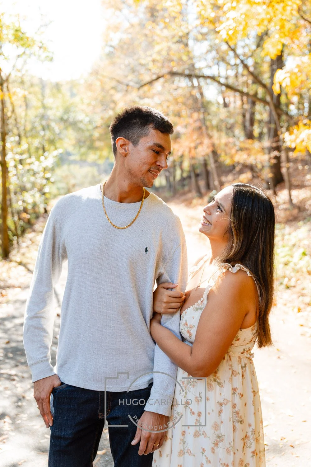 A young couple smiling and gazing at each other while standing on a sunlit autumn forest path, surrounded by trees with yellow and orange leaves.