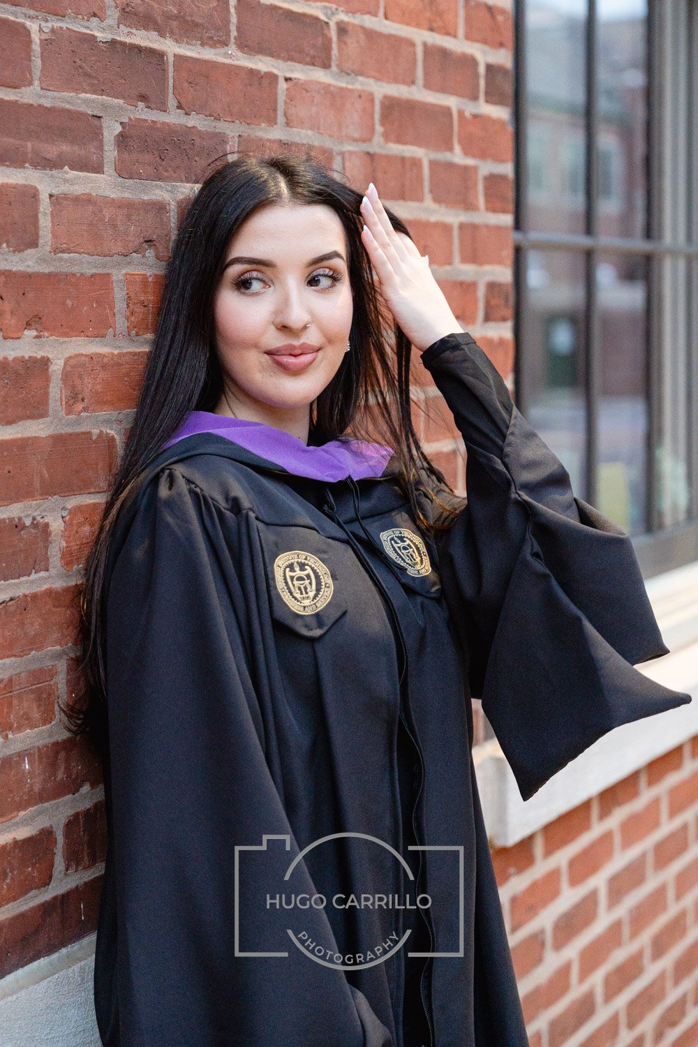 A young woman with long dark hair and fair skin wearing a black graduation gown with a purple hood, standing against a red brick wall, adjusting her hair with her right hand, looking slightly to the side.