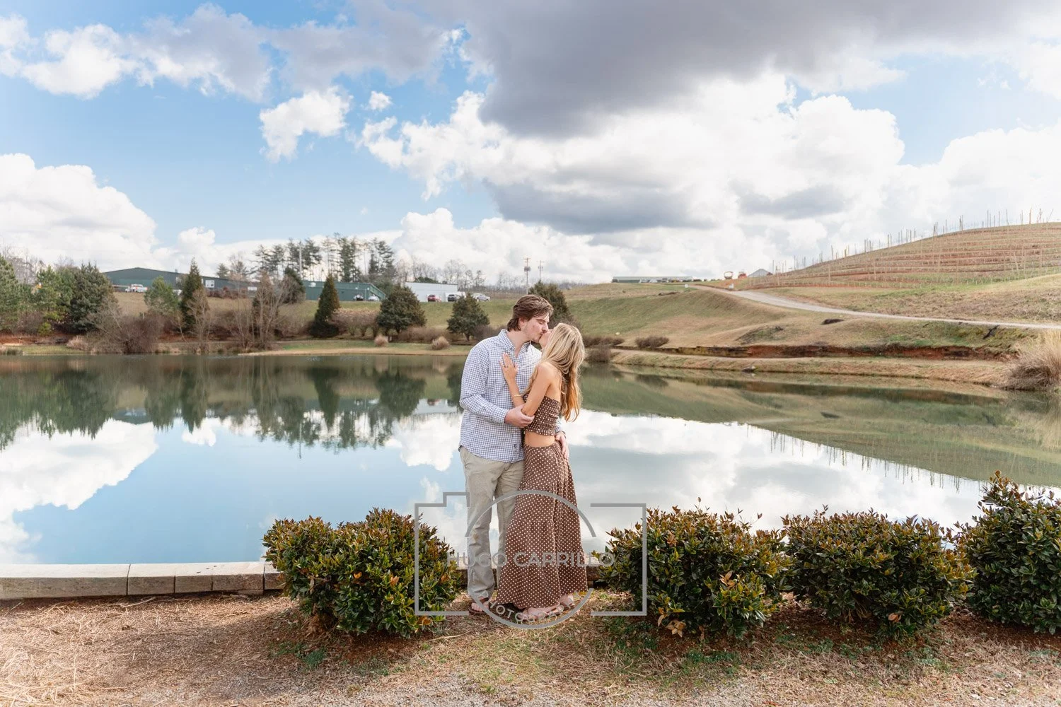 A young couple standing by a lake, sharing a kiss during daytime with partly cloudy skies and a rural landscape in the background.