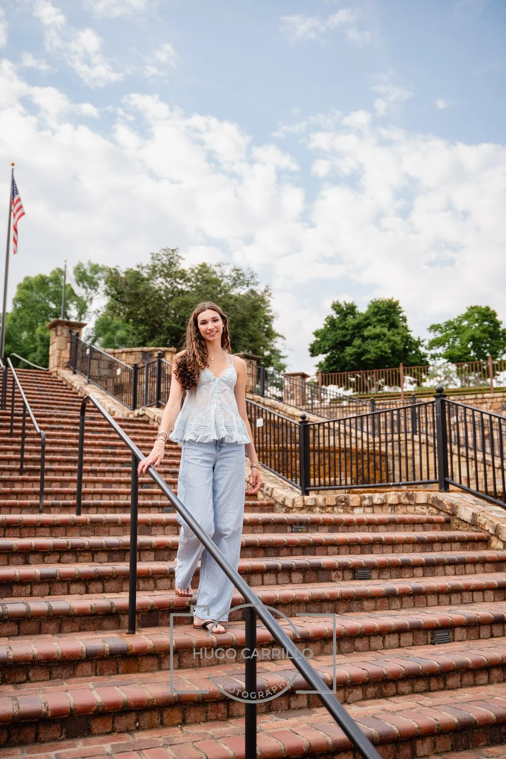 Young woman with long curly hair, wearing a light-colored sleeveless top and loose jeans, walking down a brick outdoor staircase with a metal handrail, smiling at the camera, under a partly cloudy sky with trees in the background.