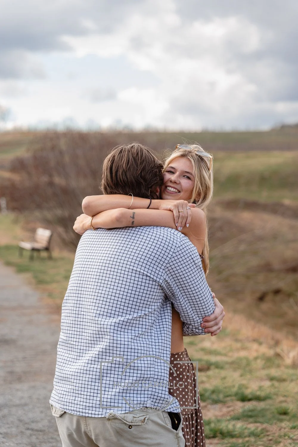 A young woman with blonde hair and sunglasses hugging a man with brown hair outdoors on a cloudy day. The woman is smiling and looking at the camera while embracing the man, who is facing away from the camera. There is a bench and grassy landscape in