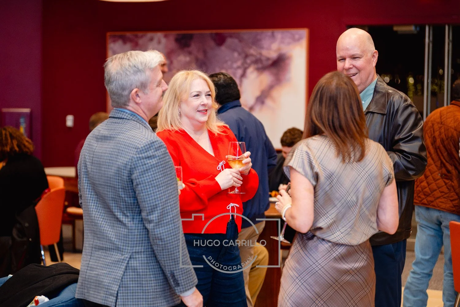 Four adults engaged in conversation at a social gathering, some holding wine glasses, in a room with red walls and artwork in the background.