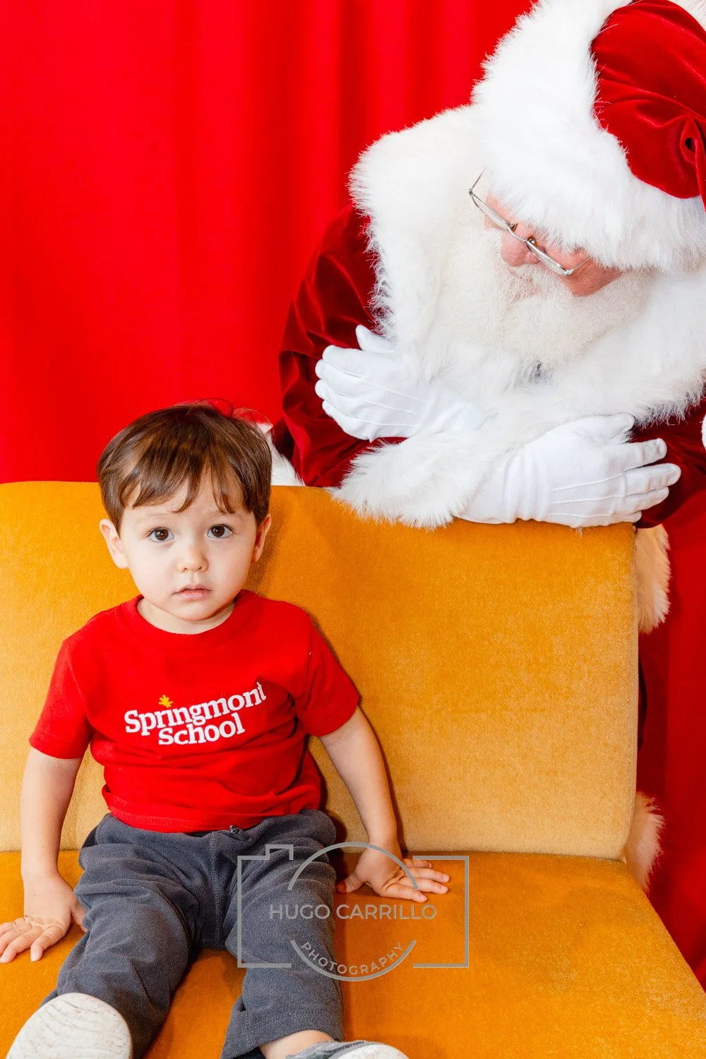 Young boy sitting on a yellow couch with Santa Claus leaning over him, both against a red background. The boy wears a red T-shirt with 'Springmont School' and gray pants.