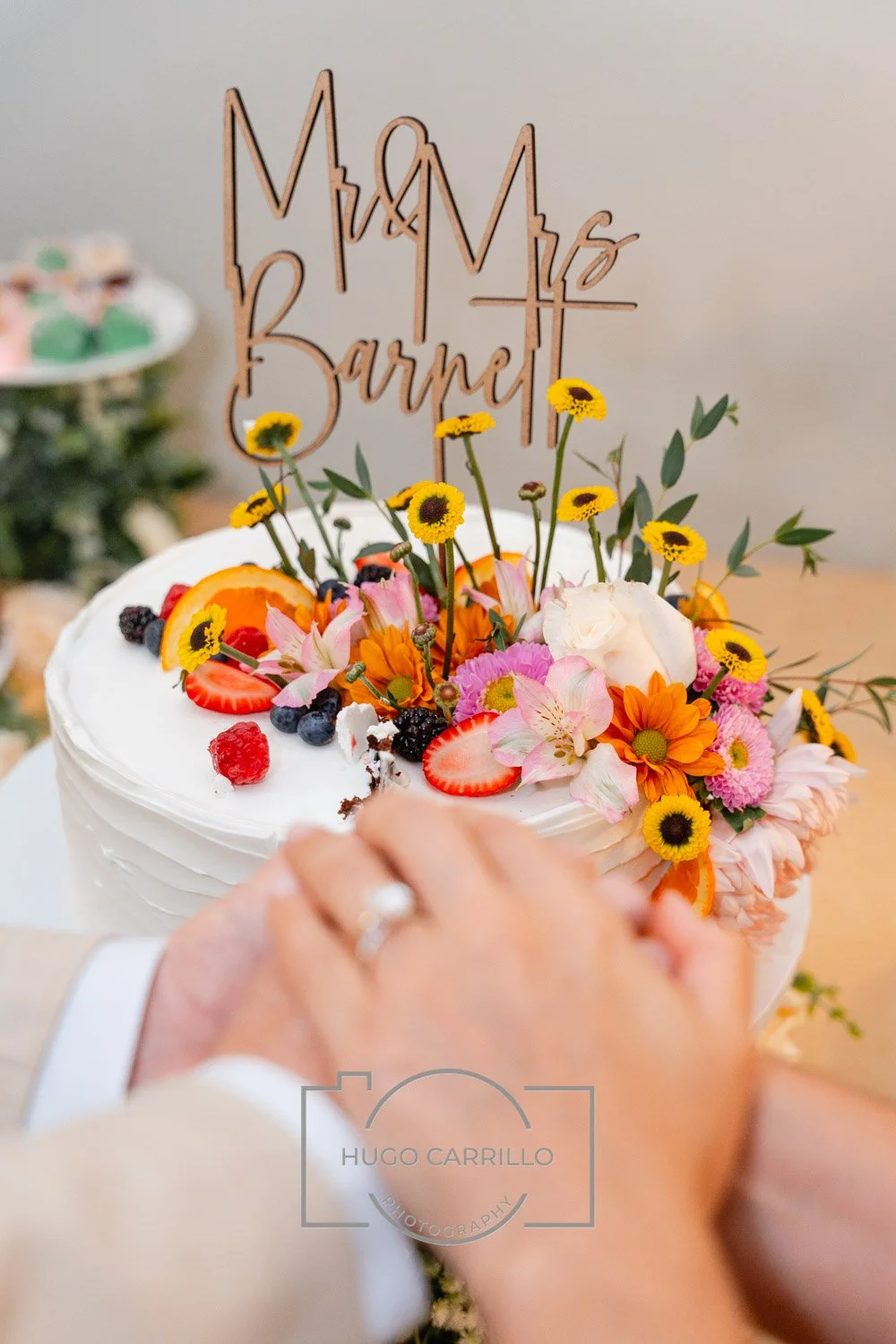A wedding cake decorated with fresh fruit and flowers, topped with a wooden cake topper reading 'Mr & Mrs Barnett', with a hand wearing a wedding ring reaching to cut it.