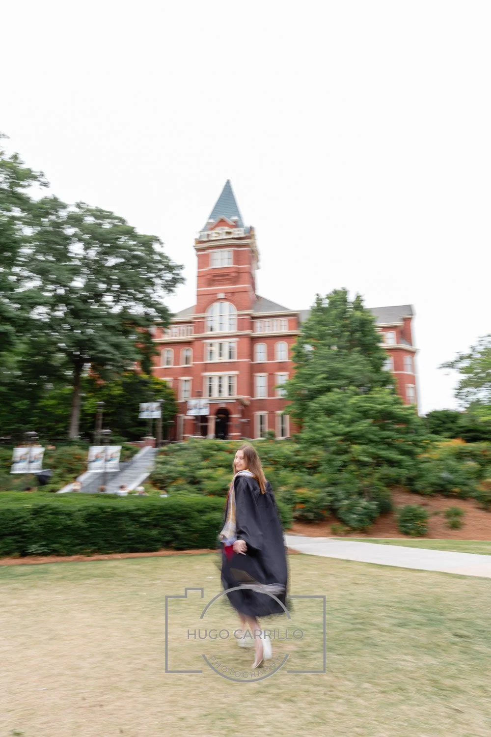 A young woman in a graduation gown walking on a grassy area near a historic red brick building with a tower, trees, and shrubbery in the background.