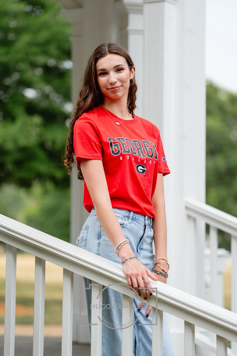 A young woman with long wavy brown hair smiling outdoors, wearing a red Georgia Bulldogs t-shirt and light blue jeans, standing beside a white railing with greenery in the background.