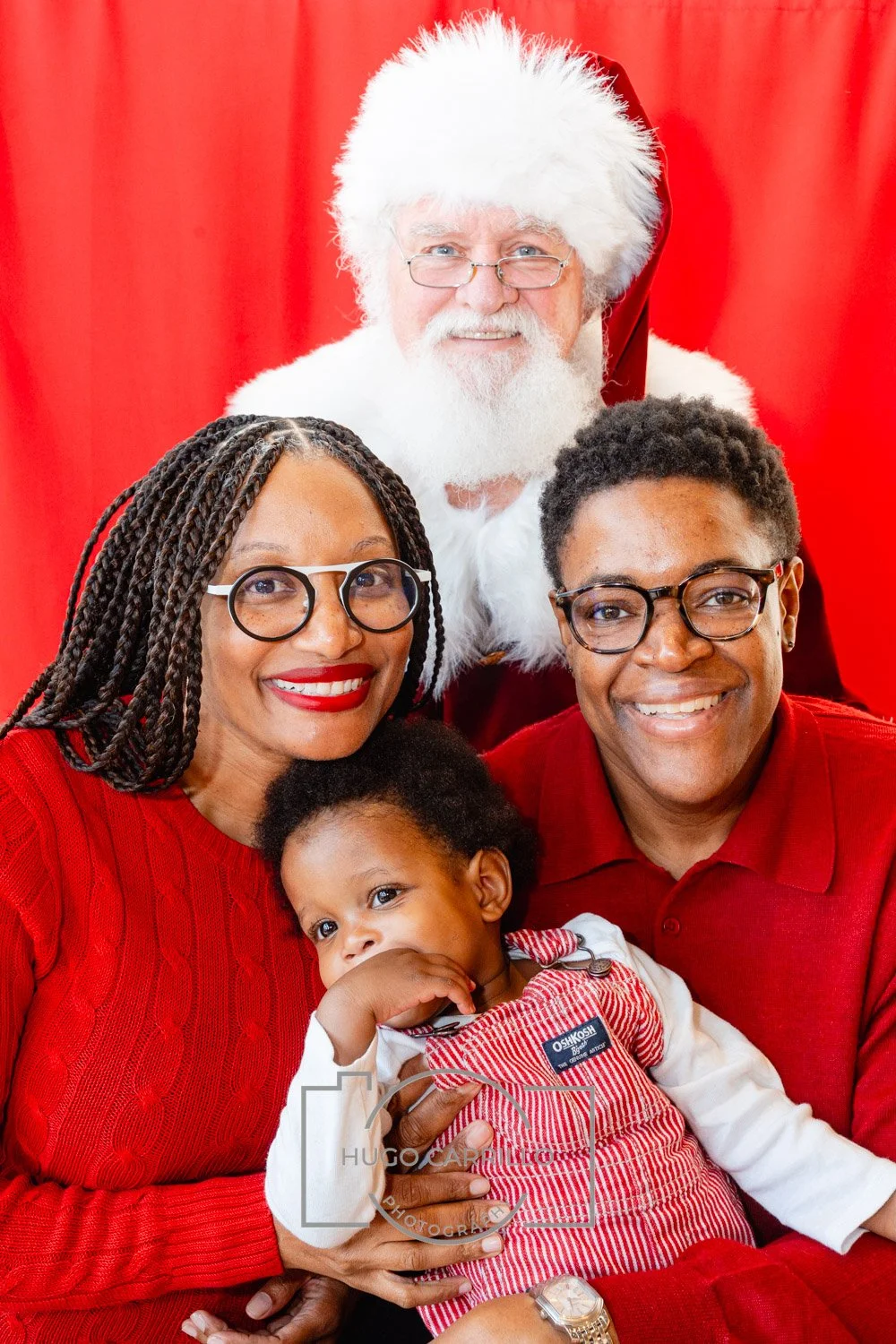 A family of four with a child, posing with Santa Claus against a red background. The woman has braids and glasses, the man has glasses, and the child is wearing a red and white striped outfit. All are smiling.