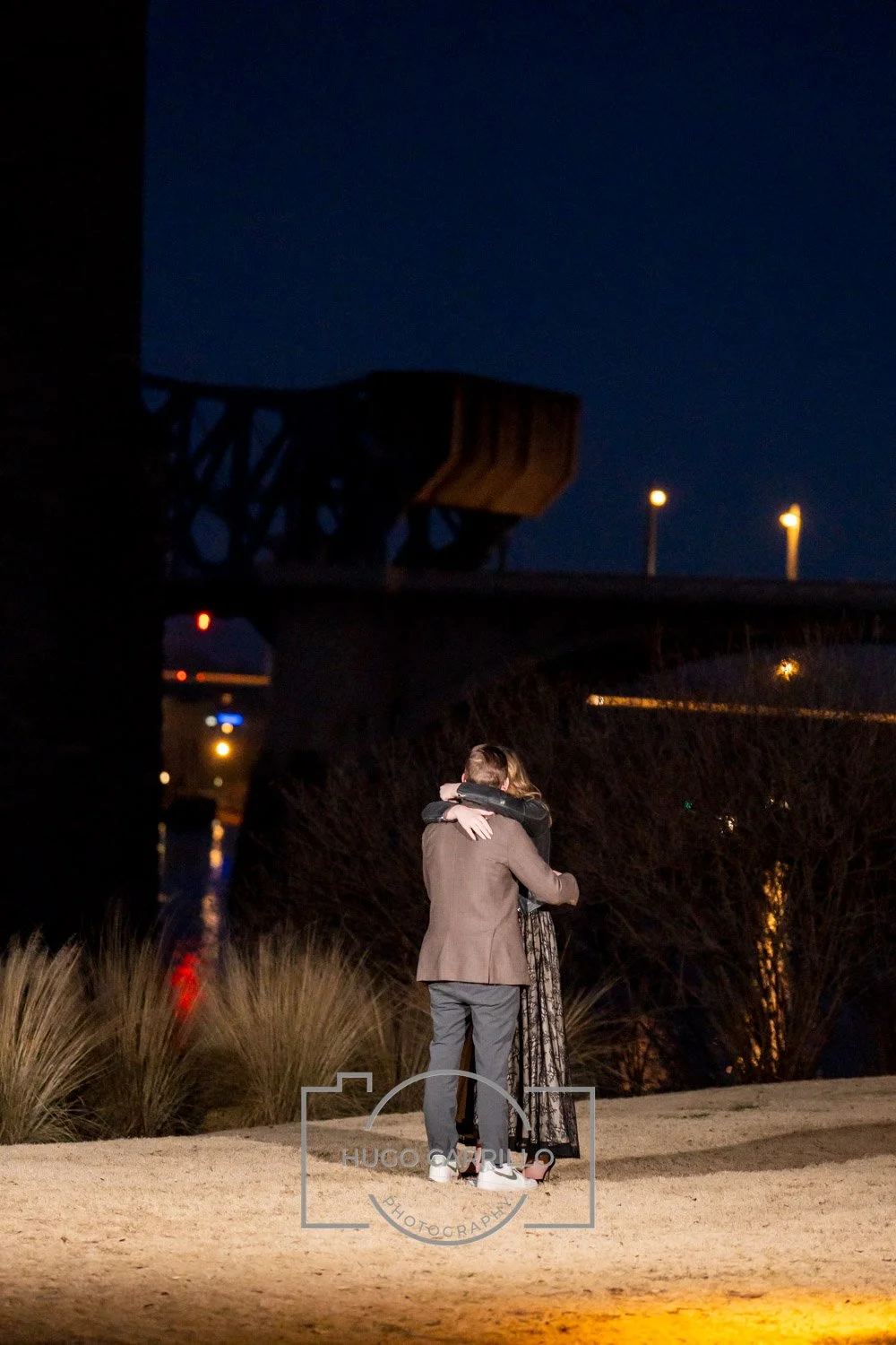 A couple embracing each other at night outdoors, with a bridge and city lights in the background.