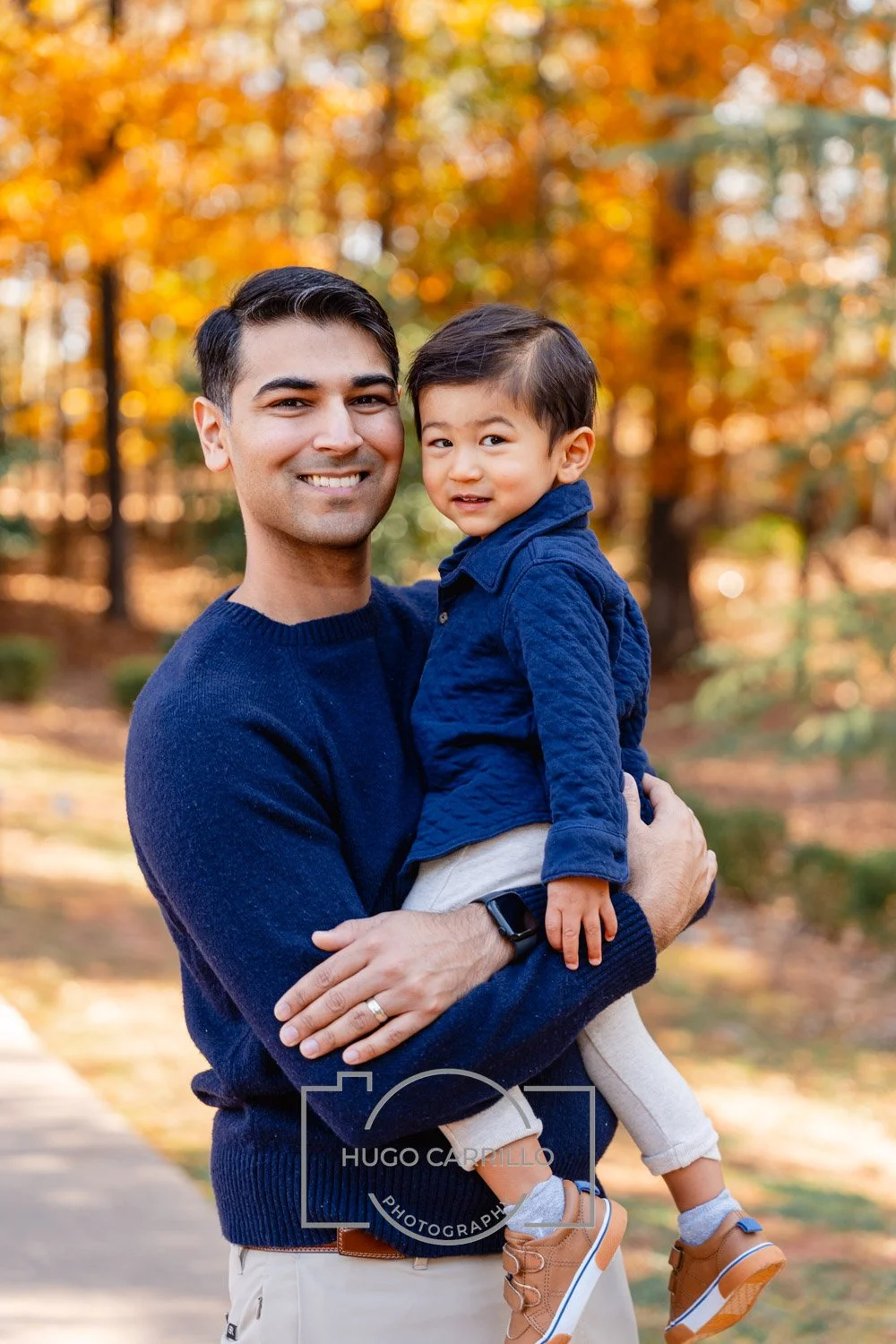 A man smiling and holding a young boy in an outdoor setting with autumn-colored trees.