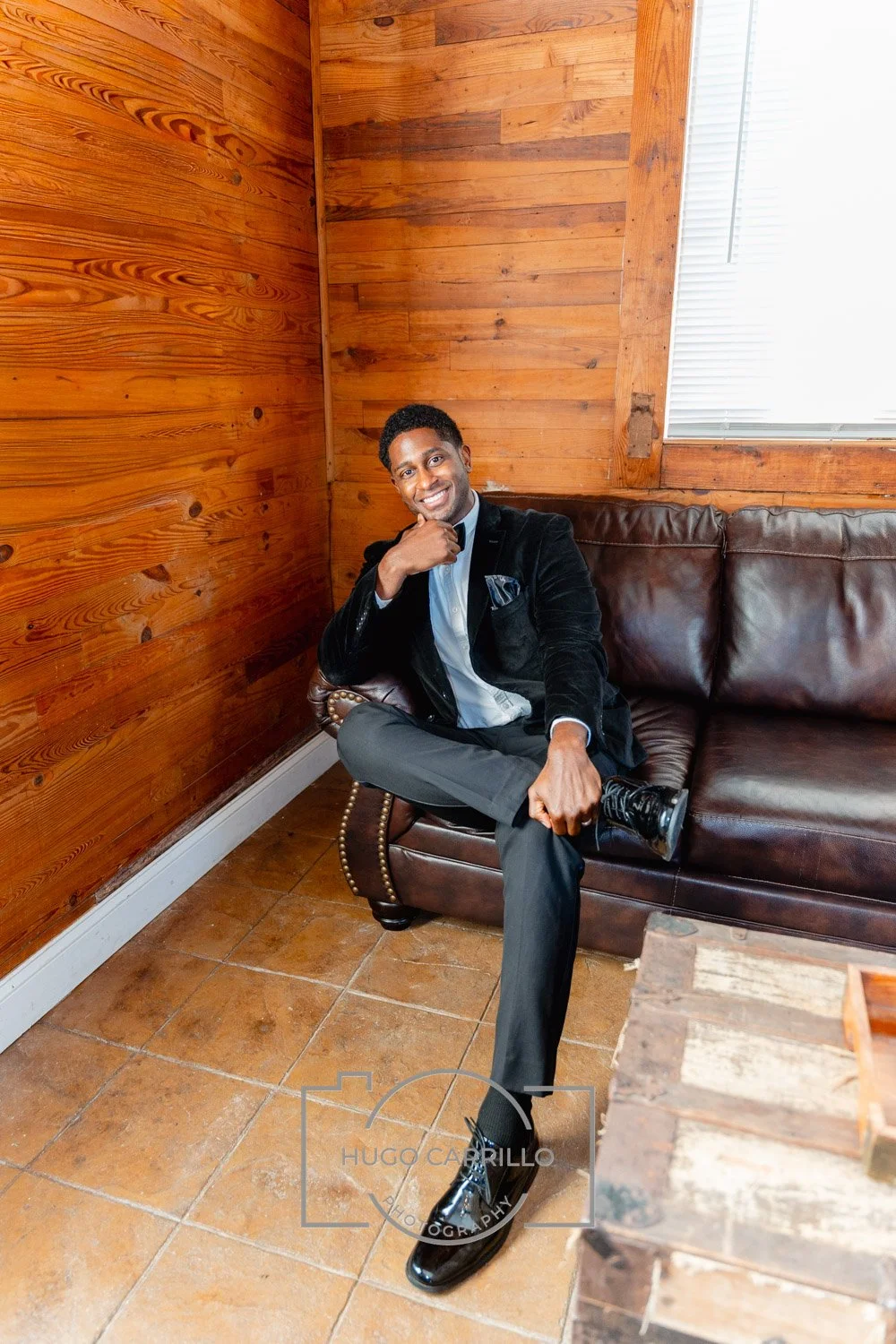 A man sitting on a brown leather couch in a wood-paneled room, smiling, with his legs crossed, wearing a black blazer, gray pants, and polished dress shoes.