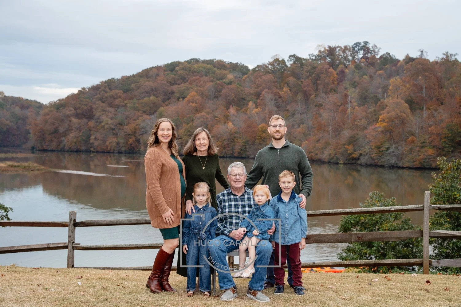 Family portrait outdoors near a lake with autumn-colored trees in the background, including three adults and four children.