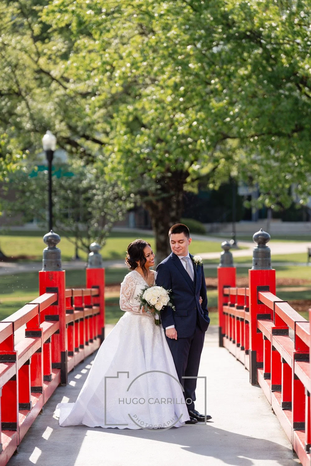 A bride and groom standing on a red bridge in a park, with trees and grass in the background, during a wedding photo shoot.