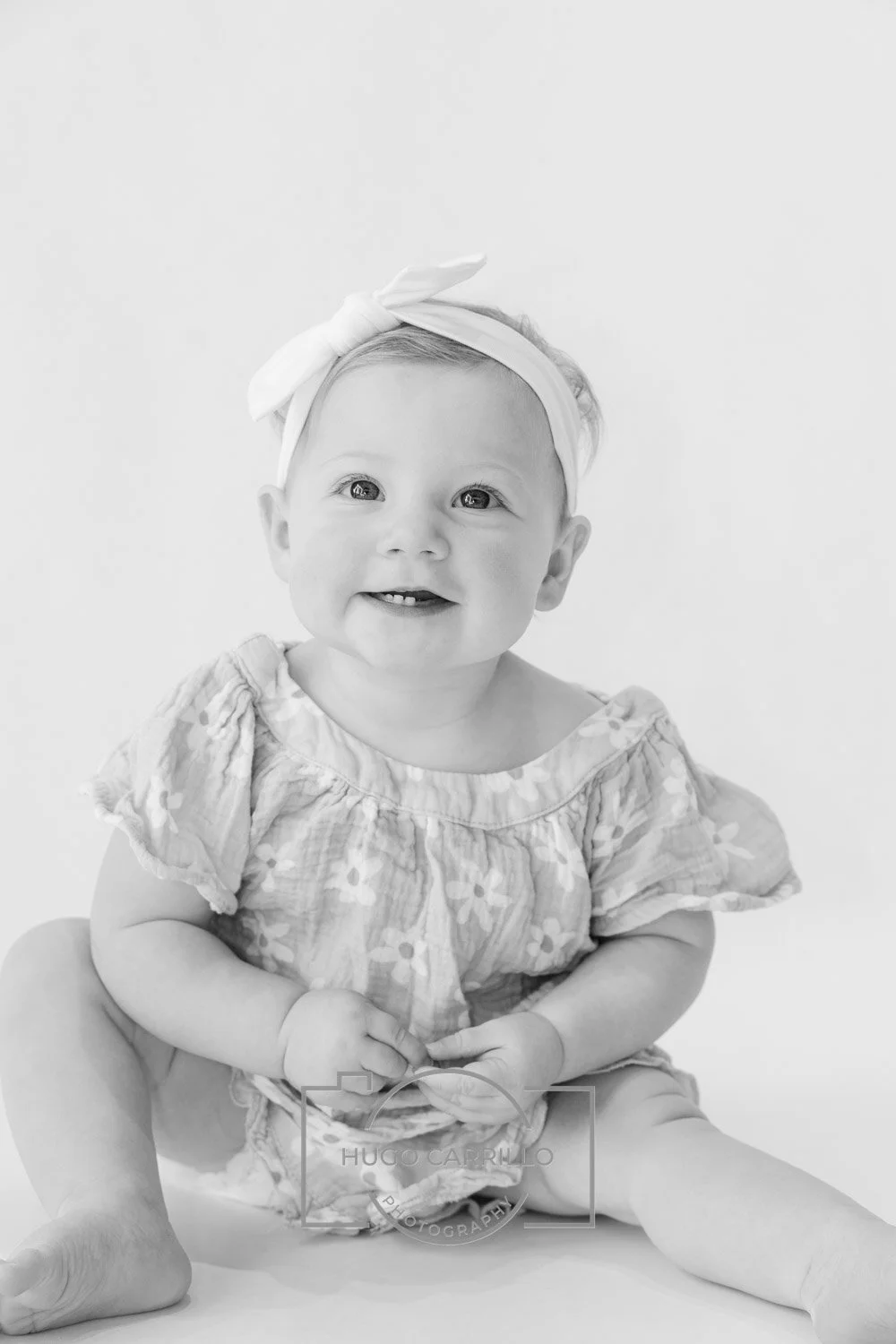 Black and white photo of a smiling baby girl sitting on the floor, wearing a floral dress and a headband with a bow.