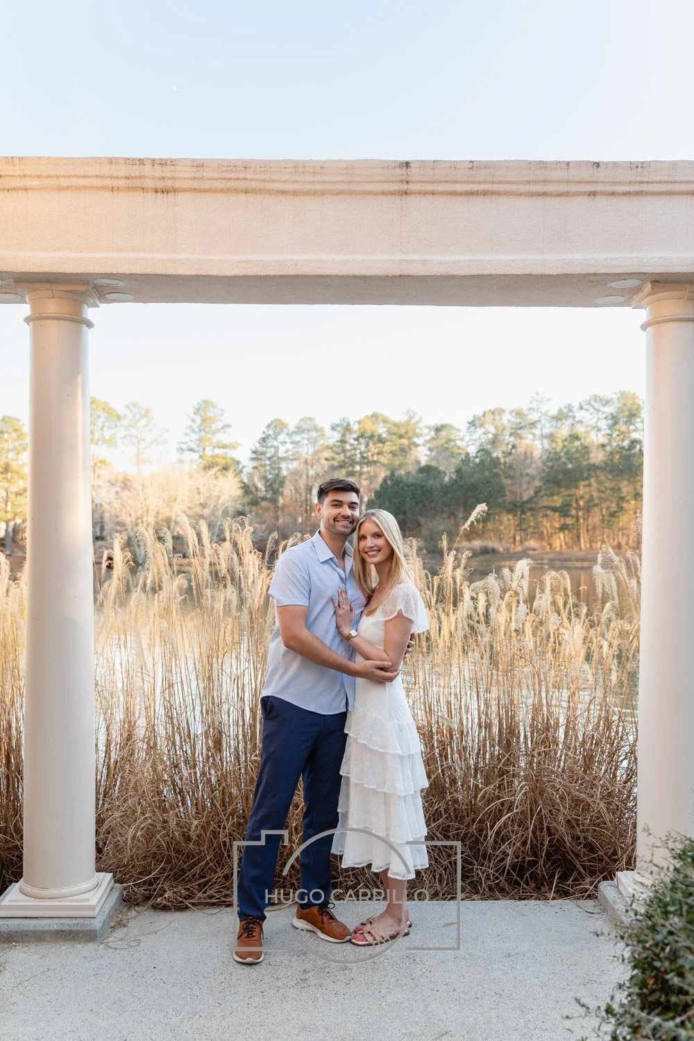A smiling couple, a man and a woman, standing close together outdoors near a pond with tall grass and trees in the background, framed by two white columns under a white archway.
