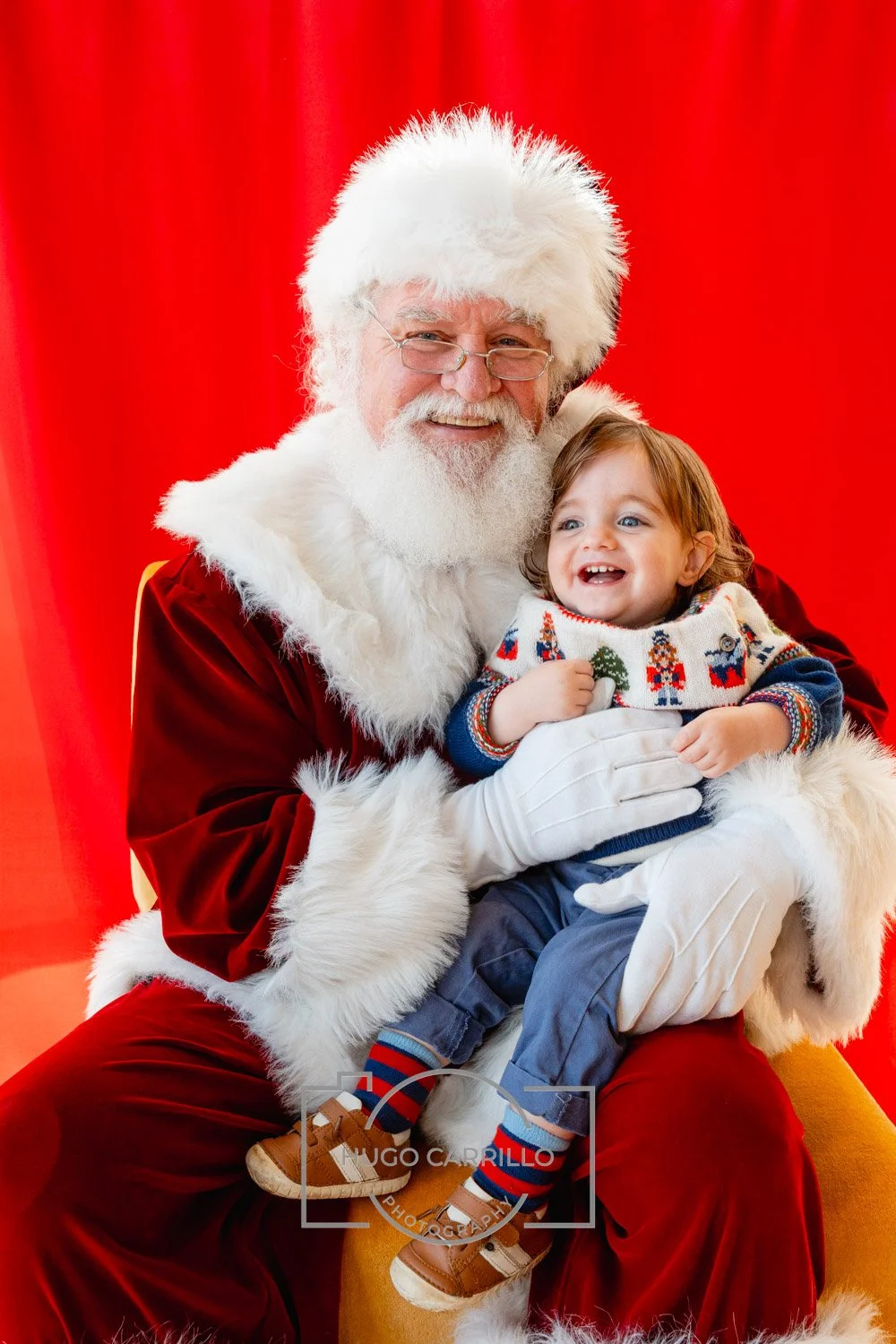 A young kid sitting on Santa Claus's lap, both smiling, with Santa wearing a red velvet suit and a white beard, against a red background.