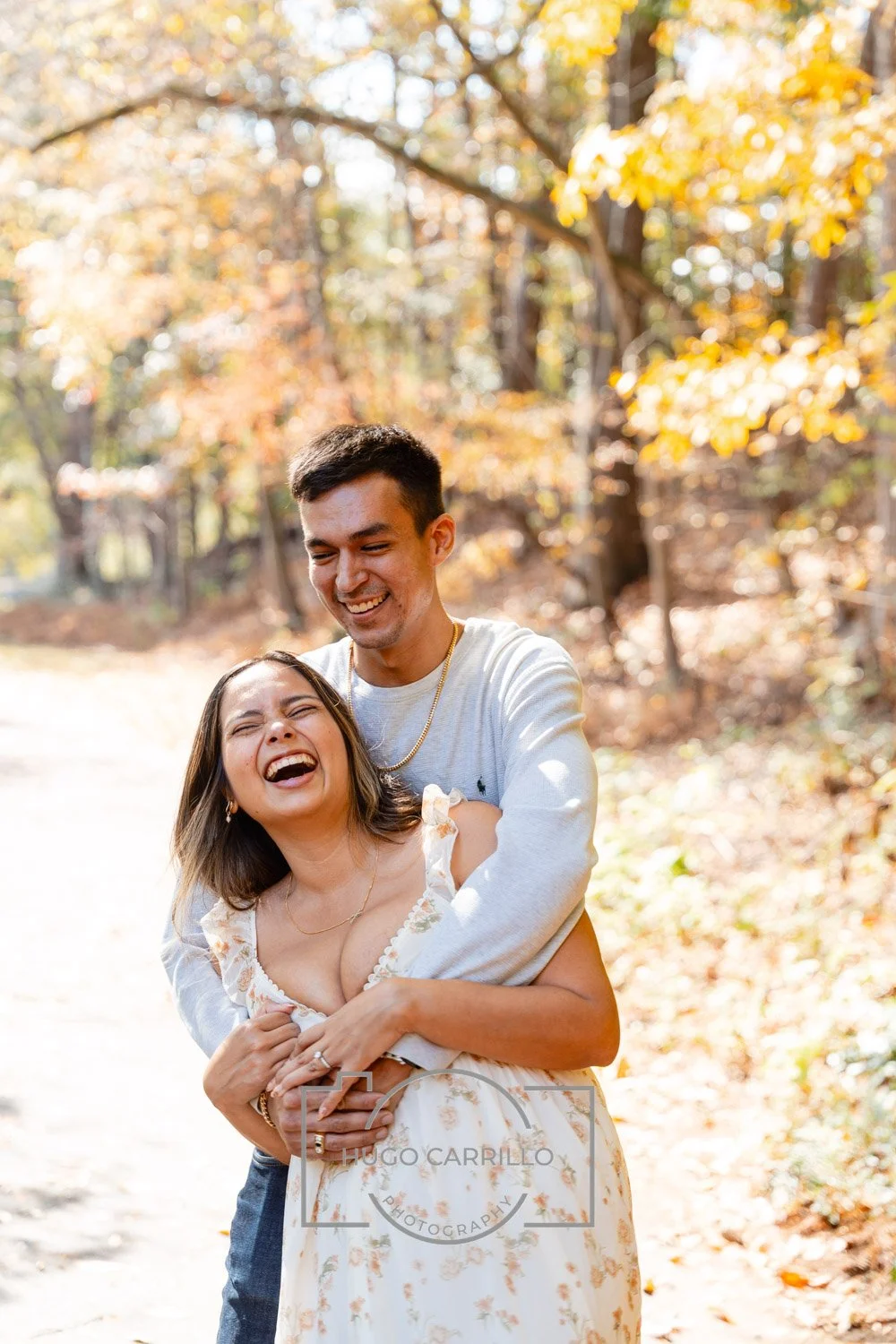 A young man and woman laughing and hugging outdoors in a park with autumn foliage.