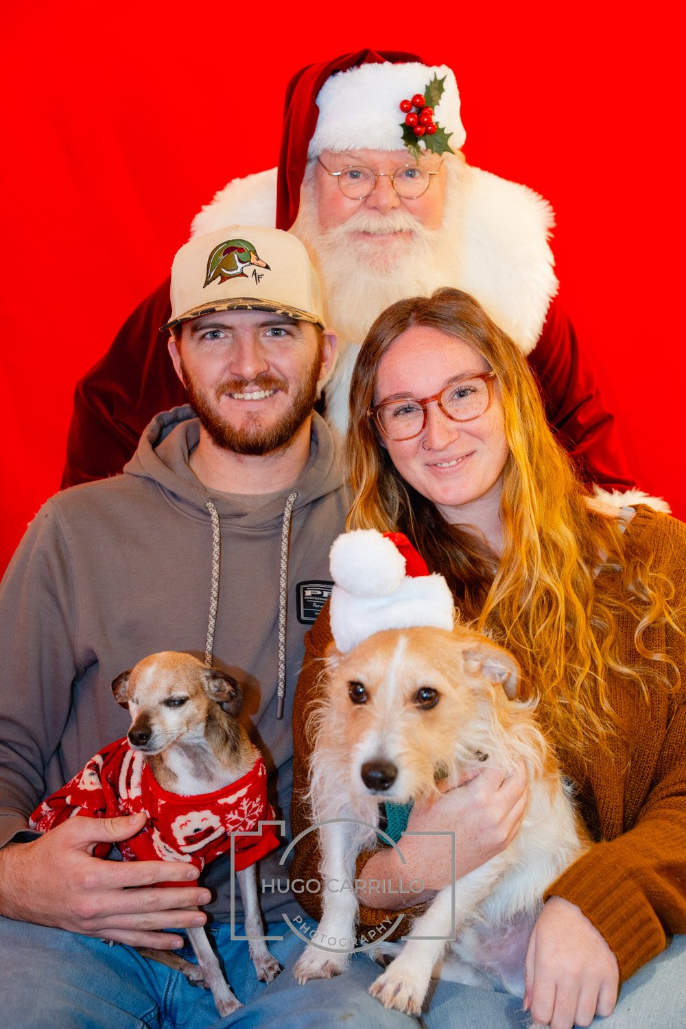 A family with two dogs poses with Santa Claus against a red background. The woman has red hair and glasses, while the man has a beard and a cap. Both dogs are dressed in holiday sweaters, and one has a Santa hat.