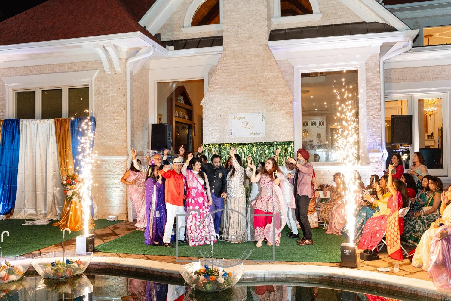 A wedding celebration taking place outdoors at night in front of a house with a lit fireplace chimney. People are dancing and celebrating on a green carpet, with sparklers and fireworks. Guests are dressed in colorful traditional attire and seated on
