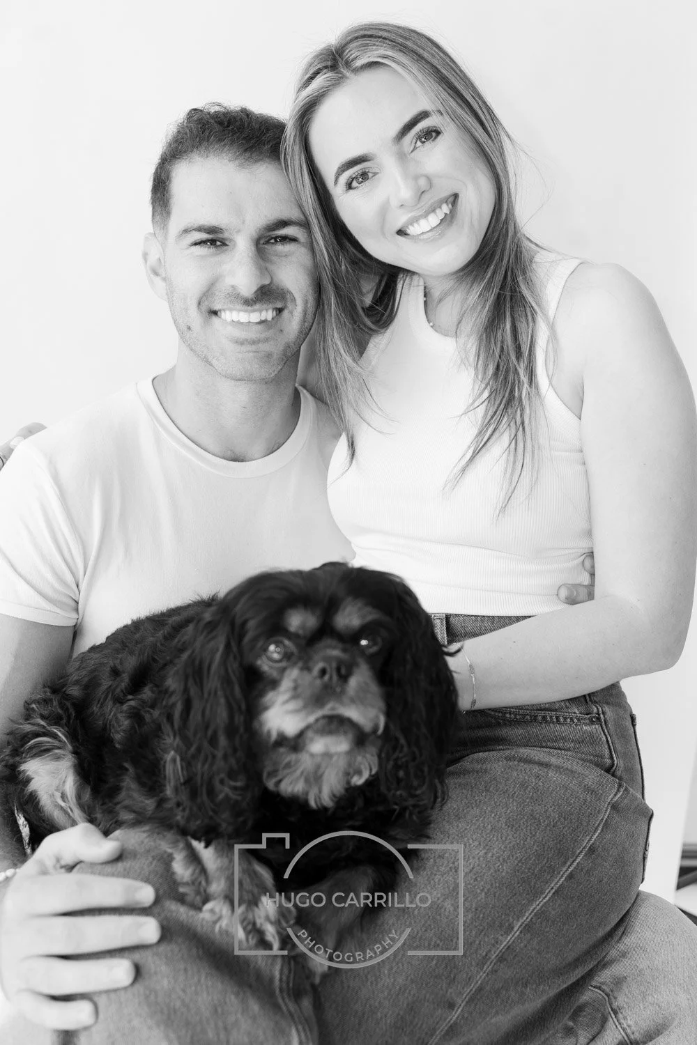 A smiling couple, a man and a woman, sitting close together with their dog, a small black and brown spaniel, in a portrait against a plain background.