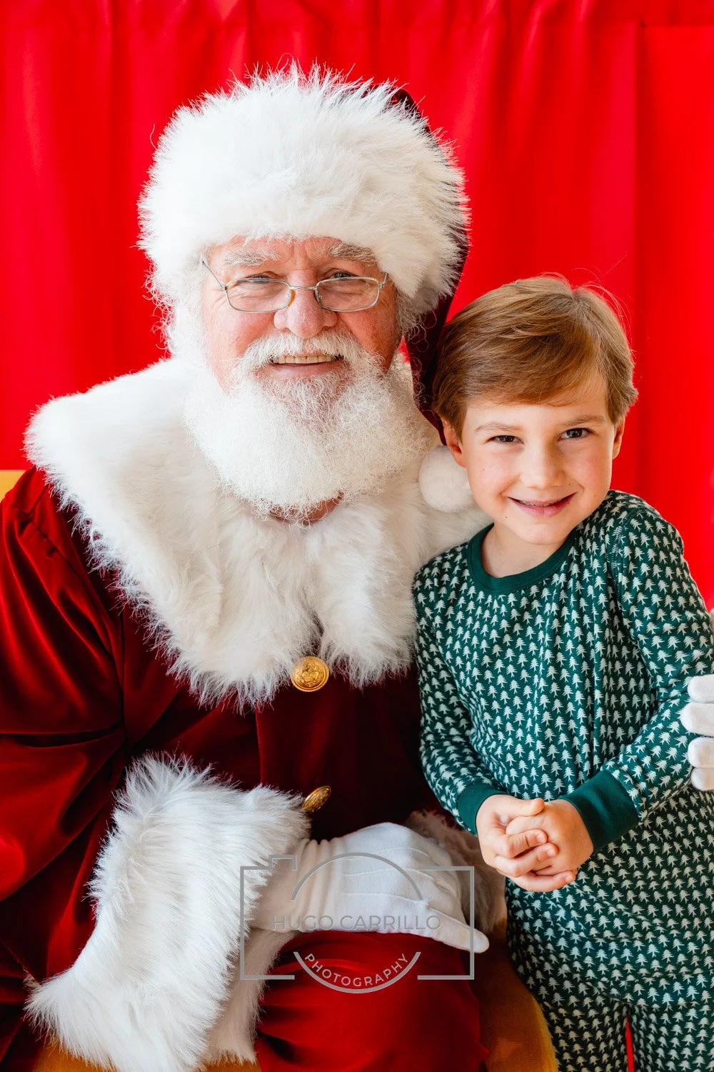 A young boy with light brown hair smiling and standing next to Santa Claus, who has a white beard, glasses, and is dressed in a red suit with white fur trim, in front of a red curtain.