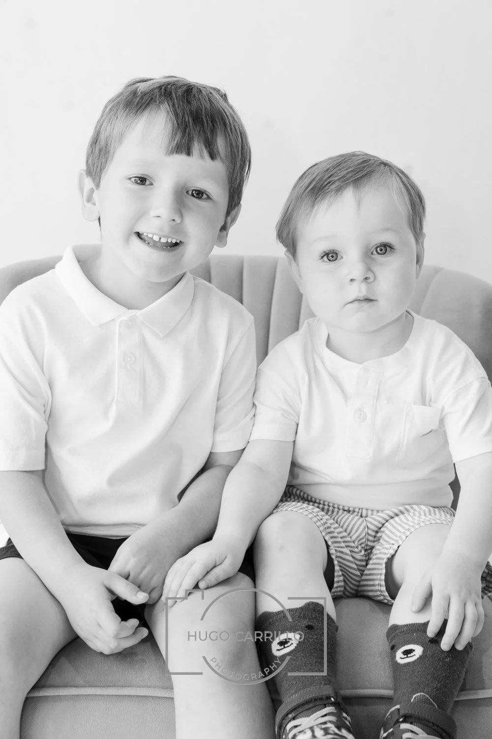 Two young boys sitting on a sofa, one smiling and the other with a serious expression, both wearing white shirts and patterned shorts, in black and white photography.