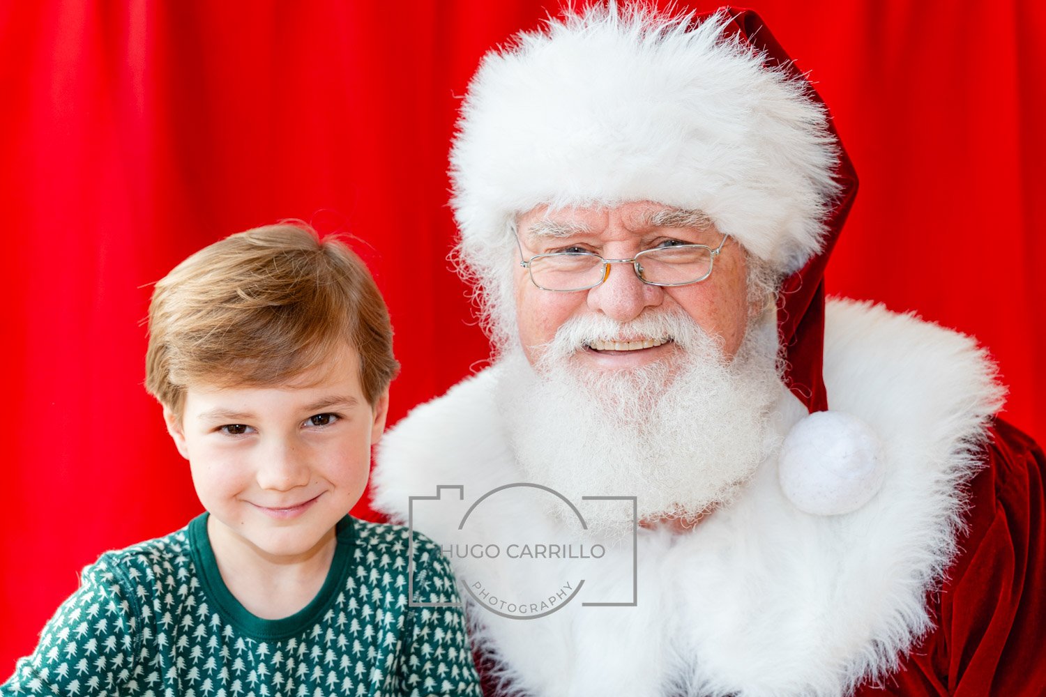 A young boy with brown hair smiling next to a man dressed as Santa Claus, who has a white beard, glasses, and a red Santa suit. They are in front of a red curtain.