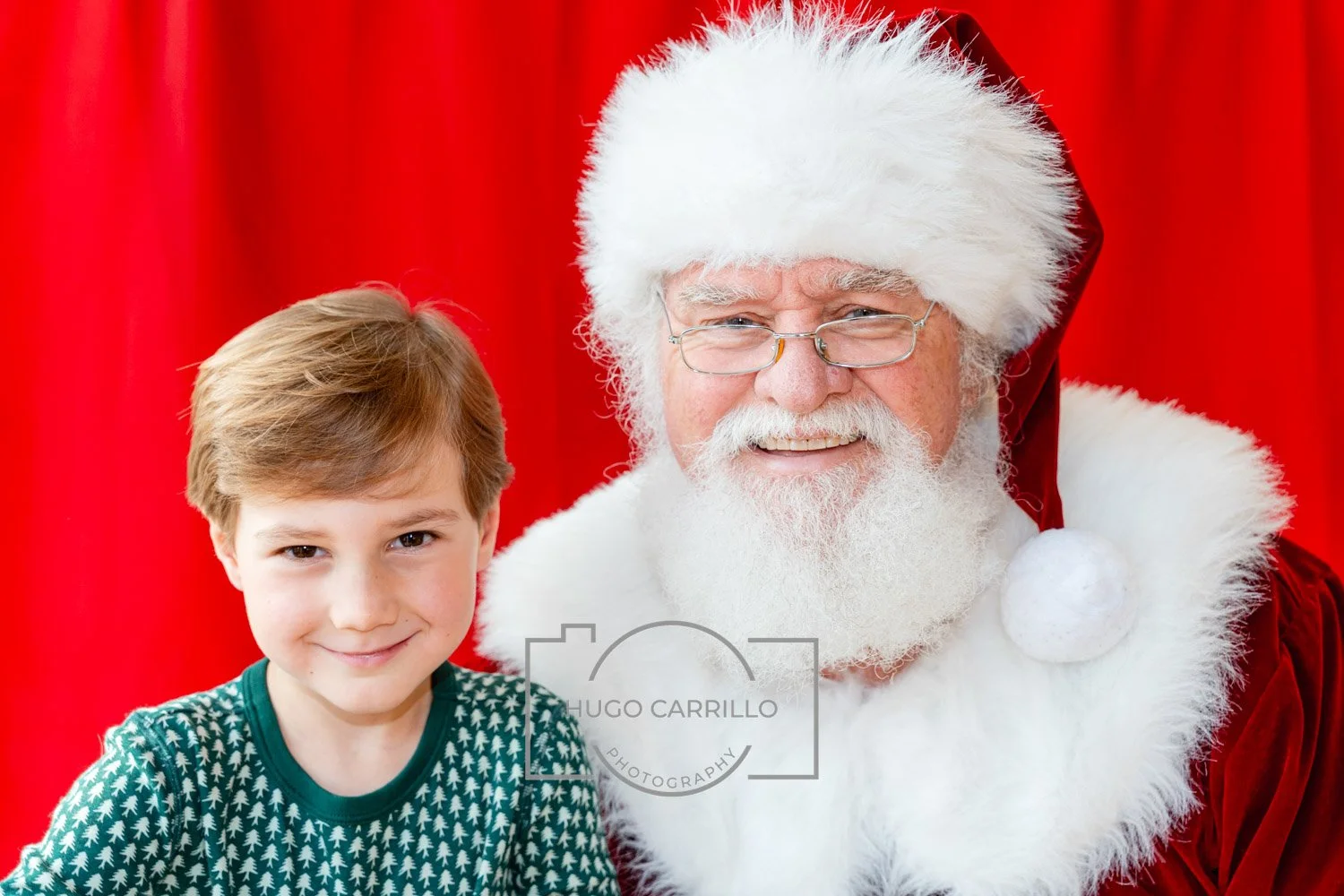A young boy with brown hair smiling next to a man dressed as Santa Claus, who has a white beard, glasses, and a red Santa suit. They are in front of a red curtain.