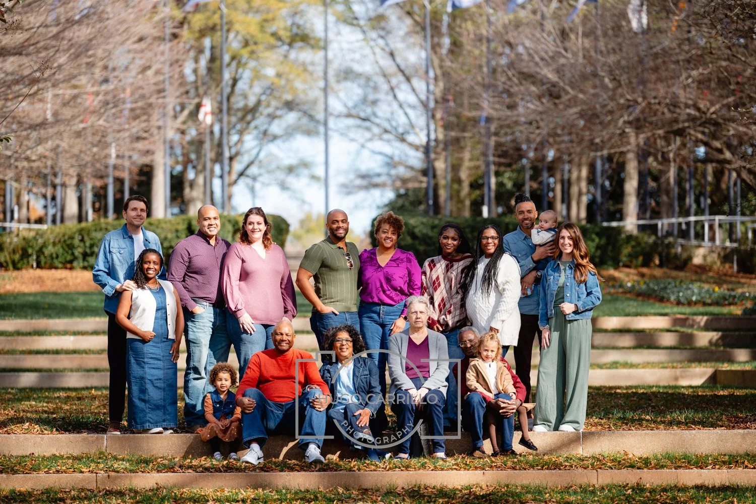 Group of people, including children and adults, posing outdoors on steps in a park during autumn with trees and fallen leaves in the background.